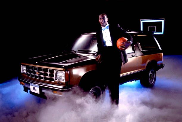 Portrait of professional basketball player Michael Jordan, of the Chicago Bulls, dressed in a tuxedo with a basketball under one arm as he poses with Chevrolet Suburban automobile, Chicago, Illinois, 1986. 